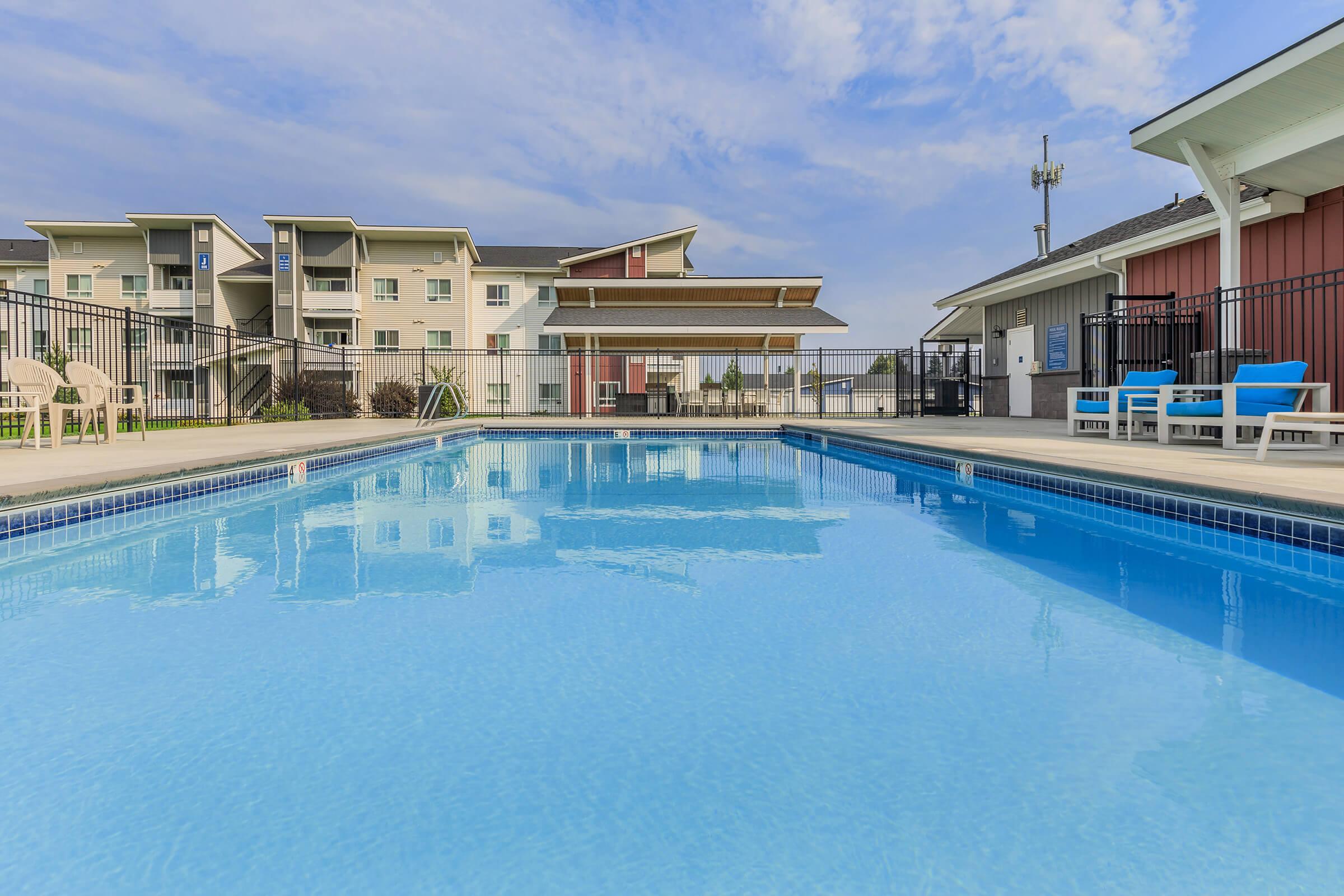 A clear swimming pool with a blue surface, surrounded by a fenced area. In the background, there are several multi-story residential buildings under a partly cloudy sky. There are lounge chairs and a shaded seating area nearby, creating a relaxing outdoor environment.
