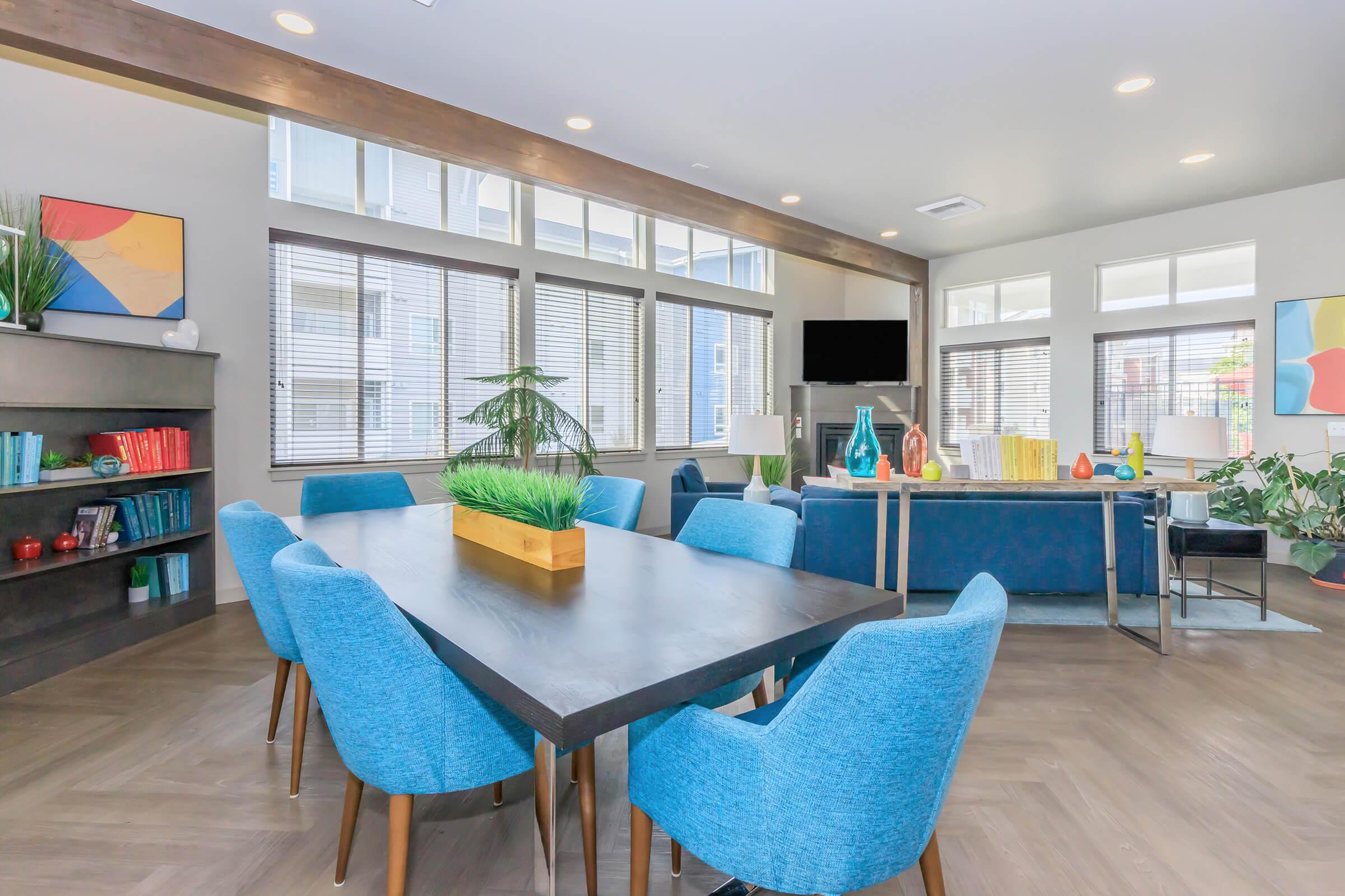 A modern communal lounge area featuring a large wooden table surrounded by bright blue chairs. There are bookshelves with decorative items, large windows allowing natural light, and a cozy seating arrangement with colorful decor and plants.