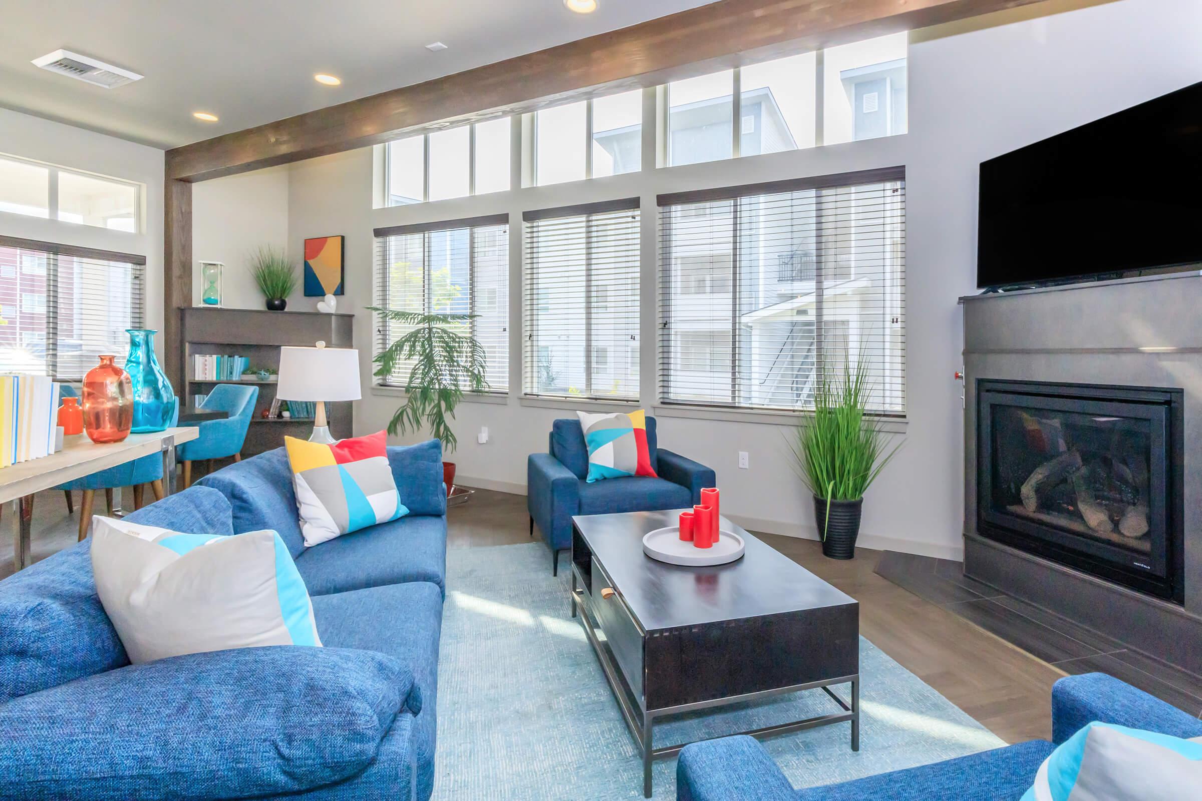 A modern living room featuring blue sofas with colorful cushions, a coffee table with decorative items, and a stylish fireplace. Bright natural light floods through large windows, enhancing the contemporary decor with plants and artwork. The overall atmosphere is inviting and cozy.