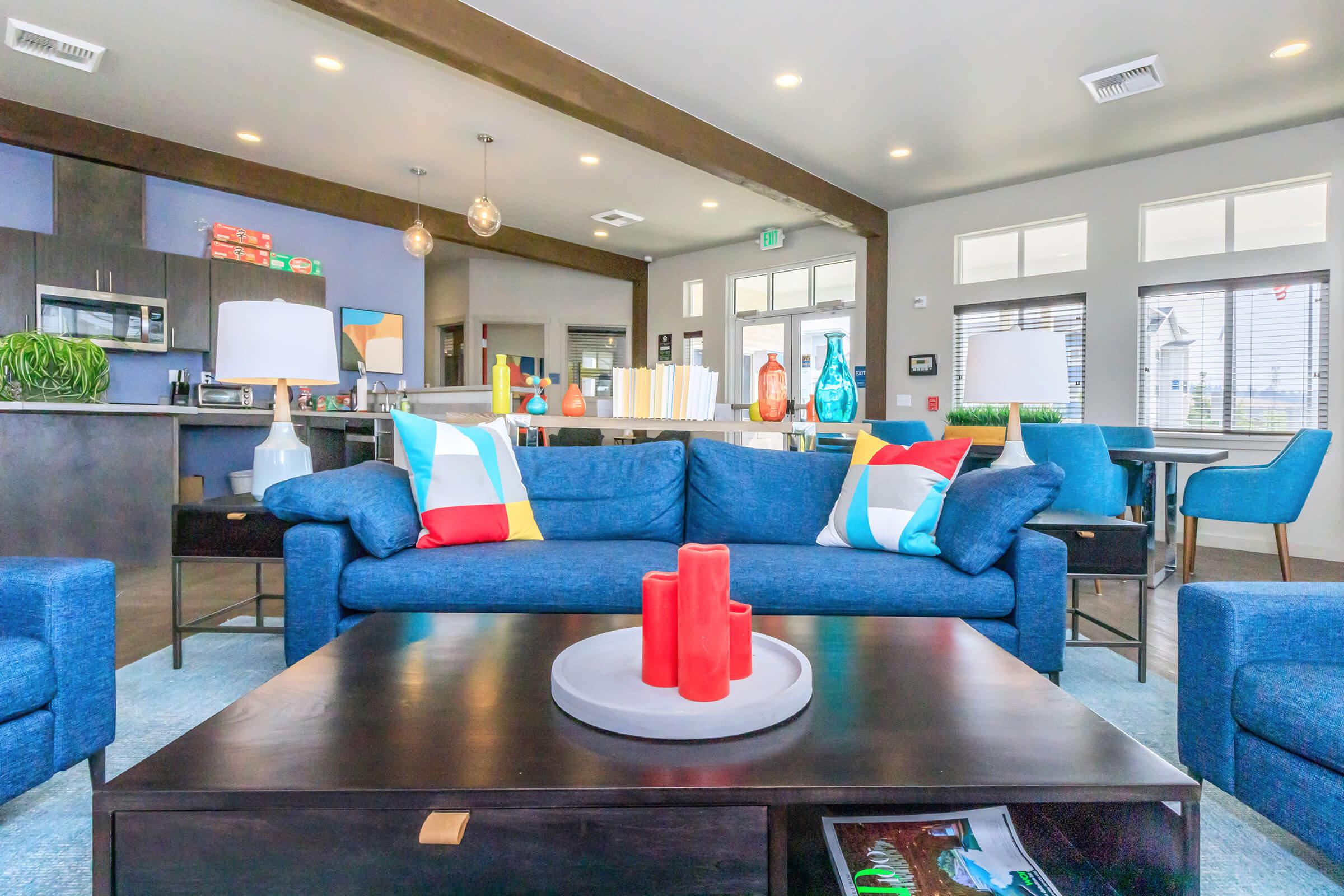 A cozy living area featuring a blue sofa with colorful cushions, a dark wooden coffee table with a round tray, and red candles. The background includes a modern kitchenette with barstools, vibrant decor, and large windows allowing natural light to fill the space, creating a welcoming atmosphere.