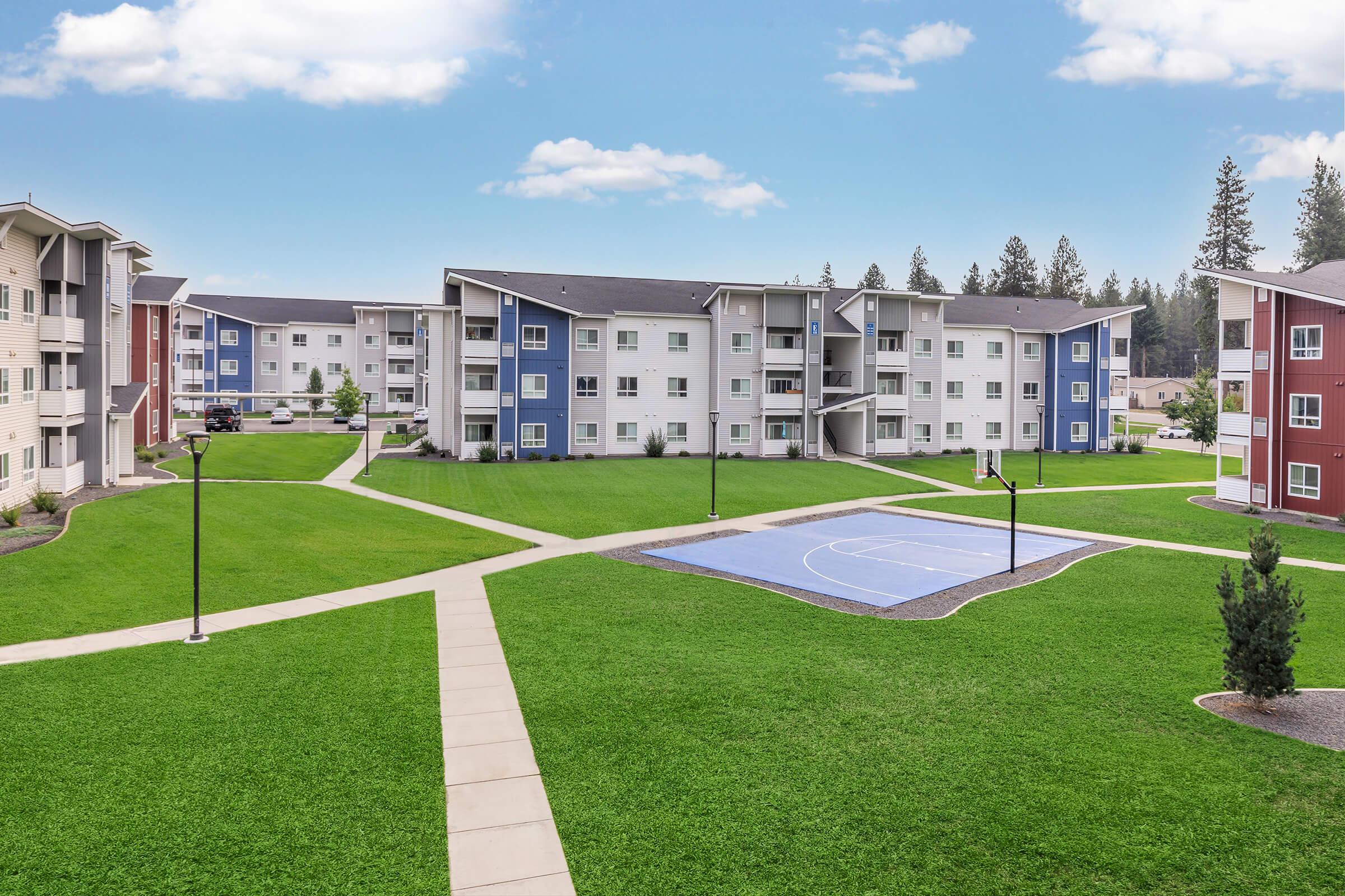 A landscape view of a residential complex featuring multiple three-story buildings in various colors. The area includes well-maintained green lawns, walking paths, and a basketball court. The sky is clear with a few clouds, creating a bright and inviting atmosphere.