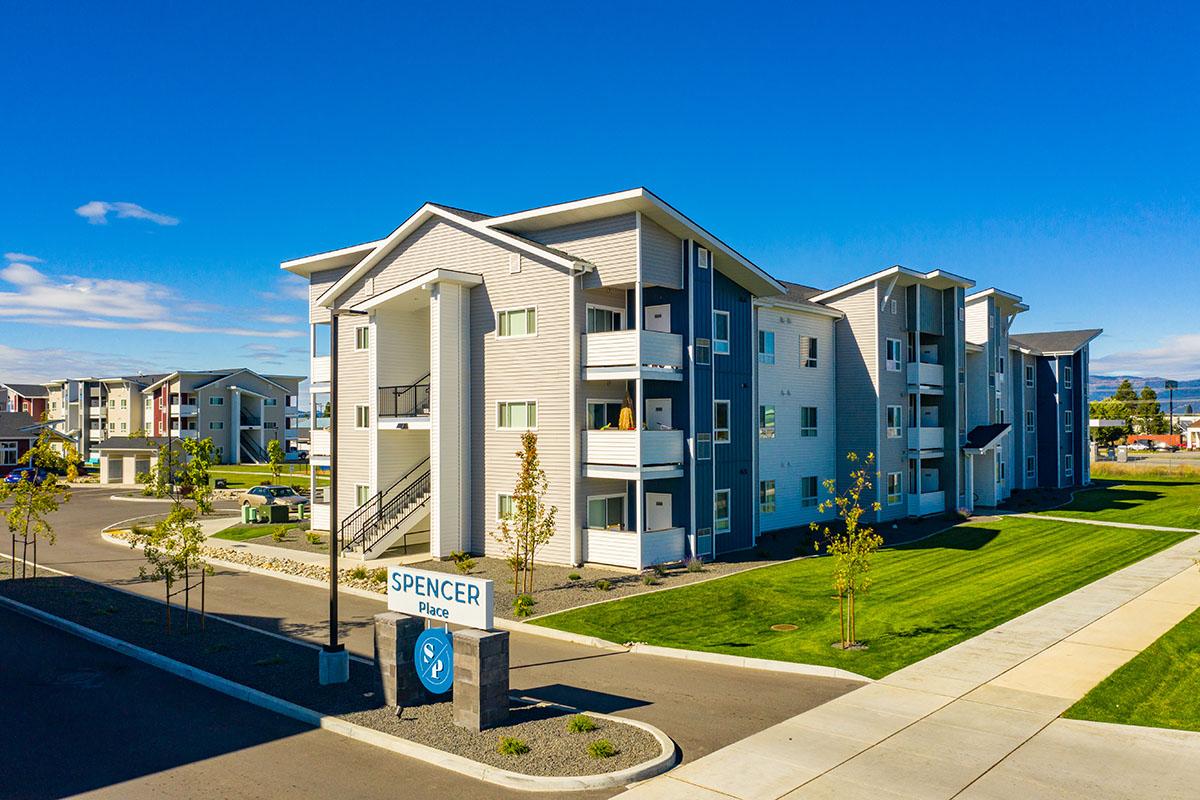 Modern apartment complex named Spencer Place, featuring multiple buildings with a sleek design, surrounded by landscaped lawns and pathways. Clear blue sky and well-maintained exterior add to the inviting atmosphere of the community.