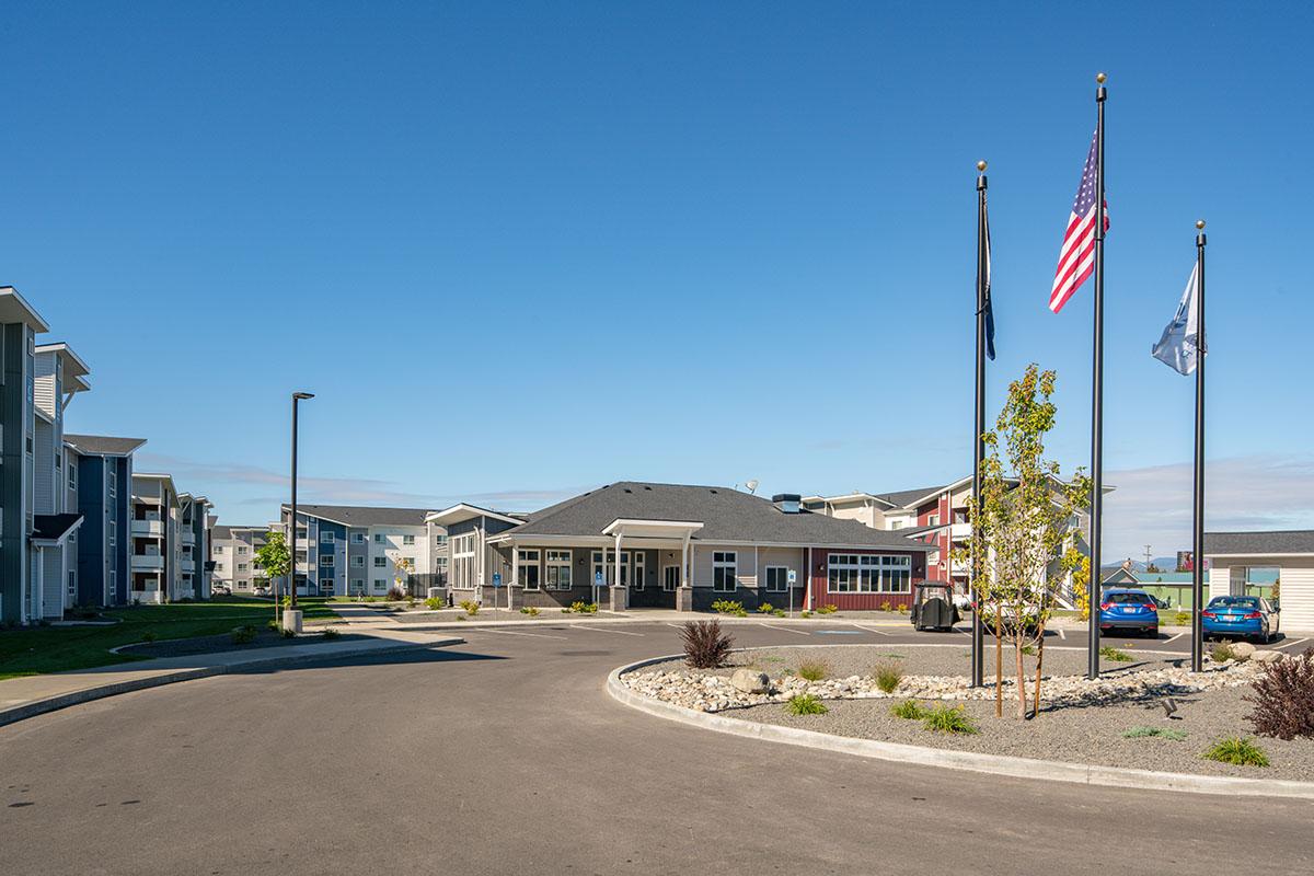 A welcoming entrance to an apartment complex featuring modern buildings, well-maintained landscaping, and three flagpoles displaying the American flag and two others. The road curves around with parking spaces visible and a clear blue sky overhead.