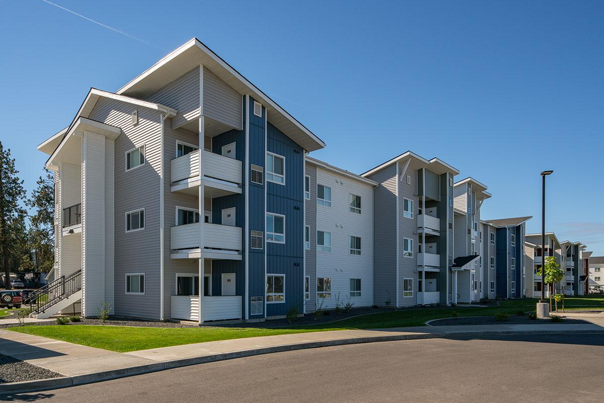 Modern apartment building with three stories featuring a combination of white and blue exterior siding. The building has multiple balconies, large windows, and a well-maintained lawn. Sidewalk and street visible in the foreground under a clear blue sky.
