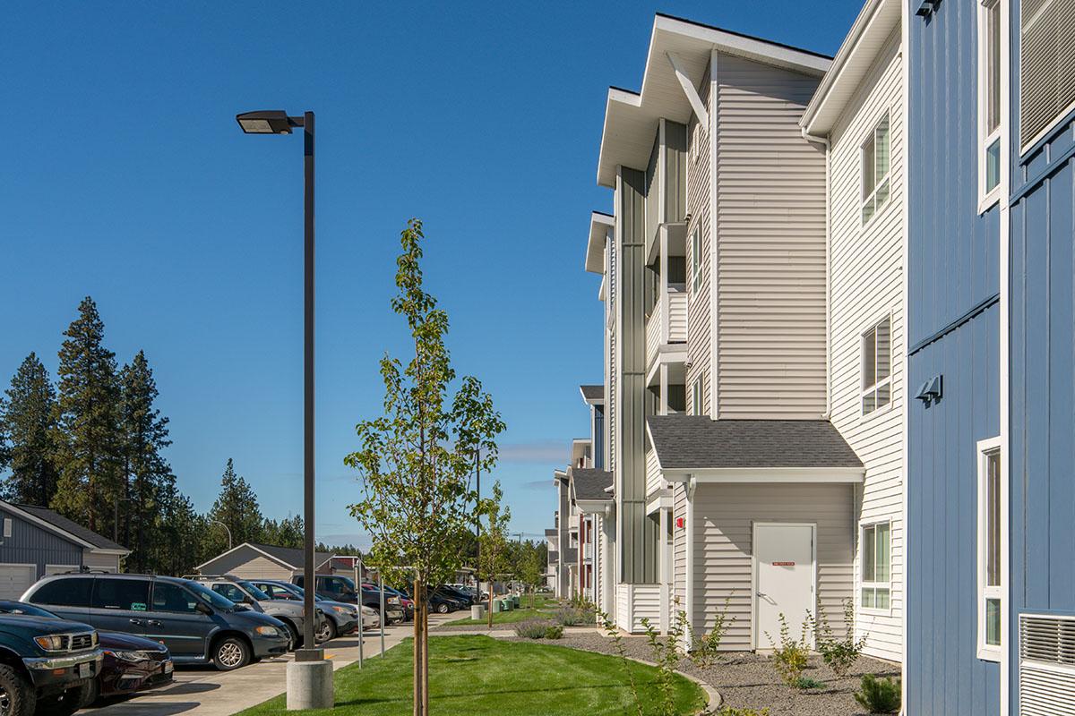 A row of modern apartment buildings with a clear blue sky overhead. The buildings feature a mix of siding colors, surrounded by well-maintained grass and small trees. Parked cars line the pathway in front, creating a suburban atmosphere. The scene conveys a sense of community and contemporary living.
