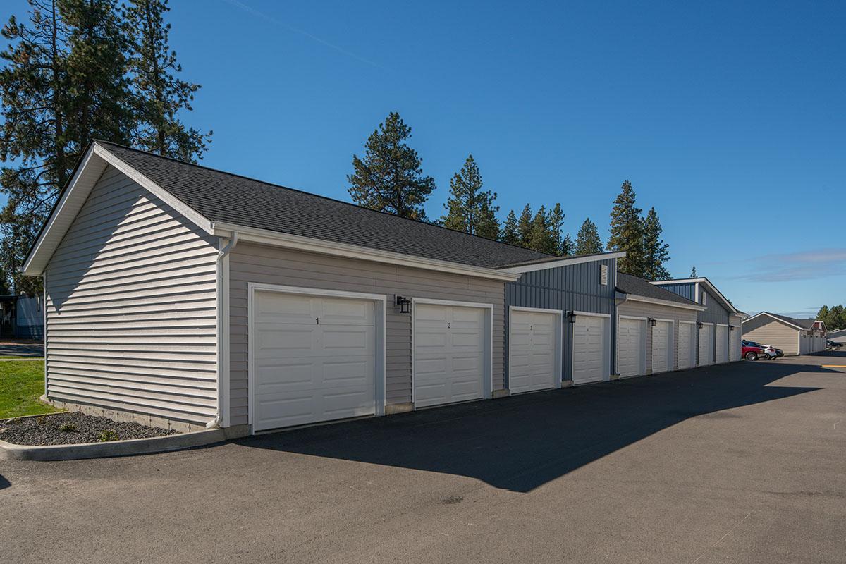 A row of garages with white doors and a gray exterior, set against a clear blue sky and surrounded by trees. The garages are situated on a paved area, indicating a well-maintained space for parking or storage.