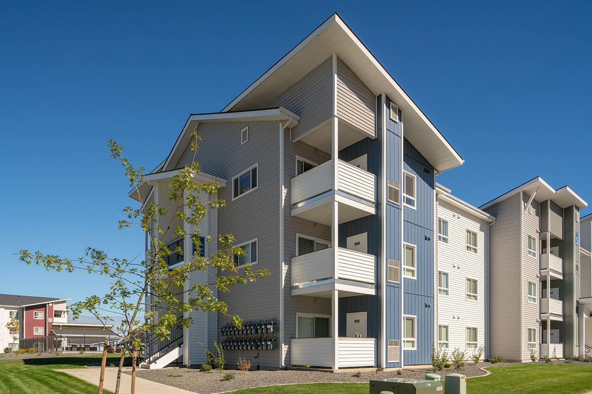 A modern multi-story apartment building featuring a combination of light gray and blue siding. The structure includes multiple balconies, large windows, and landscaped green areas in the foreground. Clear blue sky complements the overall aesthetic of the property.