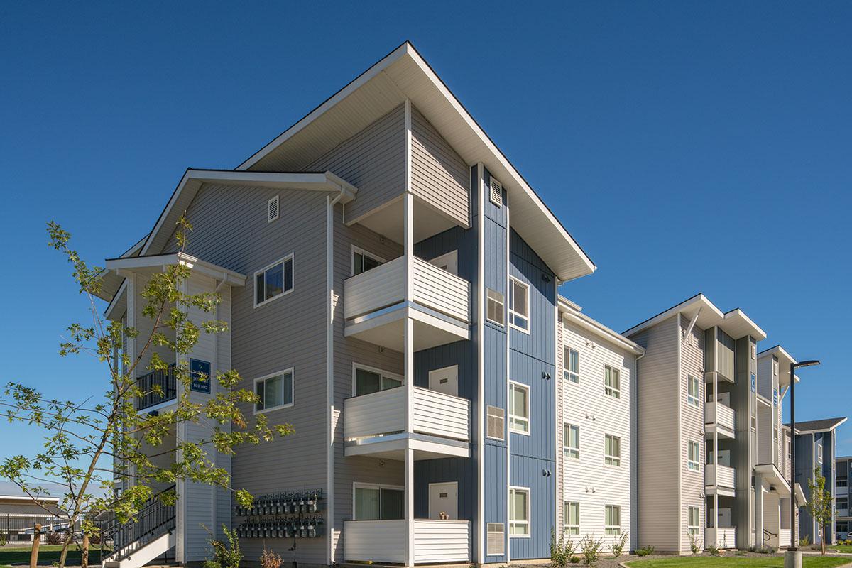 Modern apartment building with multiple stories, featuring a mix of light and dark exterior siding, balconies, and a clear blue sky in the background. Young trees are visible in the foreground, adding greenery to the scene.