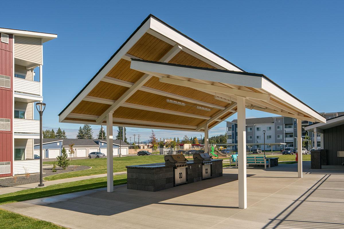 A covered outdoor pavilion with a wooden ceiling and two BBQ grills beneath. The structure is surrounded by a grassy area and nearby residential buildings. Clear blue sky in the background. Ideal for gatherings or outdoor cooking.
