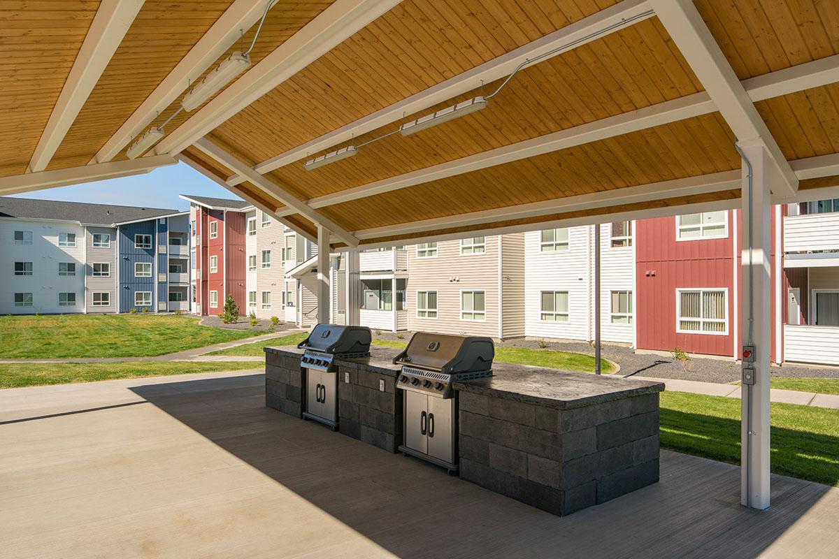 A covered outdoor grill area featuring two stainless steel BBQ grills on a stone base. In the background, several colorful apartment buildings with large windows are visible, set in a green lawn area under a clear blue sky. The space is designed for communal cooking and dining.