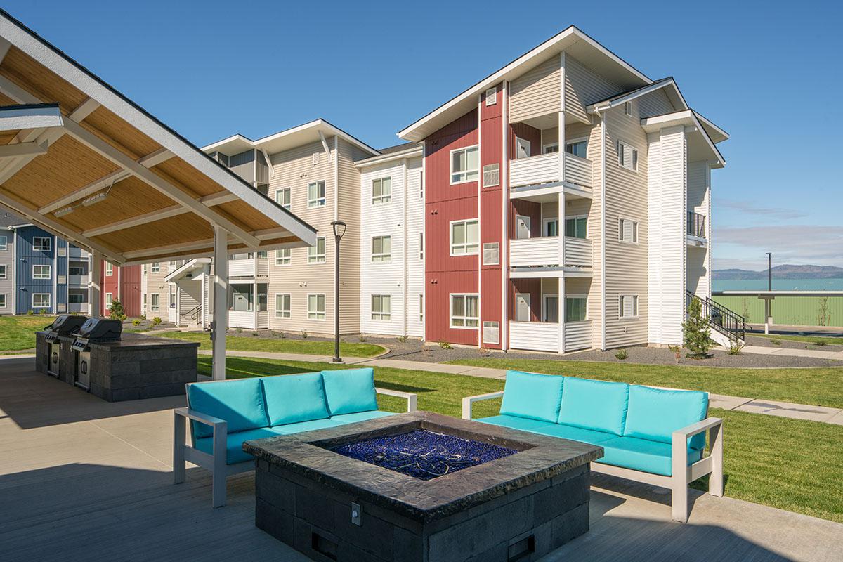 Modern apartment complex featuring multi-story buildings with a mix of white and red exteriors. In the foreground, a stylish outdoor seating area with turquoise sofas surrounds a fire pit with a blue glass surface. Grills are available under a covered patio, set against a green lawn and clear blue sky.
