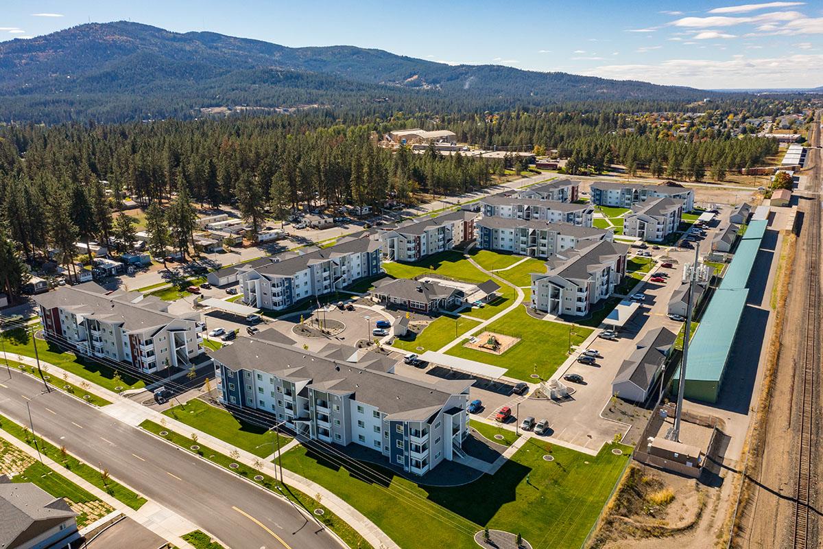 Aerial view of a residential complex featuring multiple apartment buildings arranged around green lawns and walkways, surrounded by trees and mountains in the background. The area includes streets, parking spaces, and nearby infrastructure, showcasing a suburban environment.