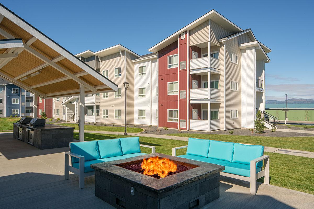 Outdoor seating area featuring turquoise couches around a fire pit with flames. Modern multi-story residential buildings in the background, set in a landscaped environment under a clear blue sky. Grilling stations are visible, enhancing the inviting atmosphere of the space.