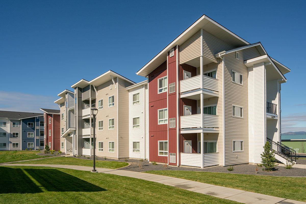 A modern multi-story apartment building featuring a mix of colors, including red and beige. The structure has several balconies, large windows, and is surrounded by a well-maintained lawn and pathway. Clear blue sky in the background enhances the bright atmosphere of the scene.