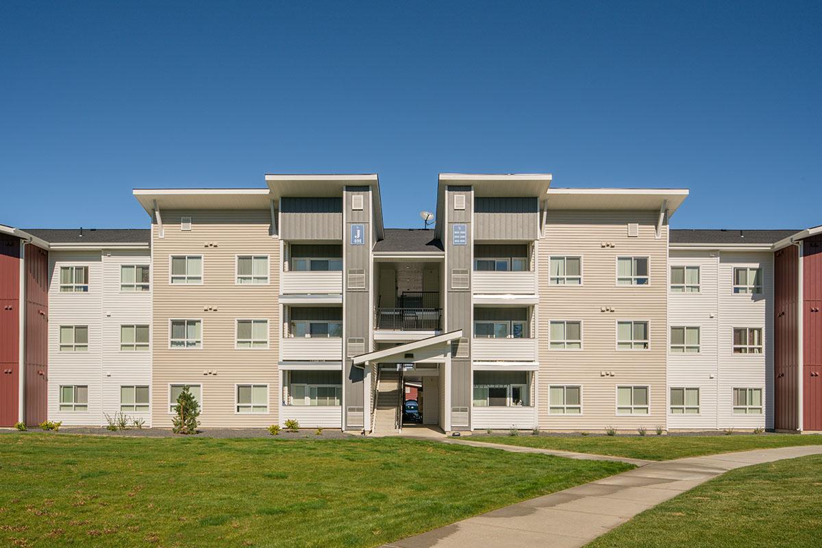 A modern three-story apartment building with beige and red exterior. It features multiple balconies, large windows, and a central entrance with a covered awning. A well-maintained lawn surrounds the structure, and a paved path leads to the entrance under a clear blue sky.