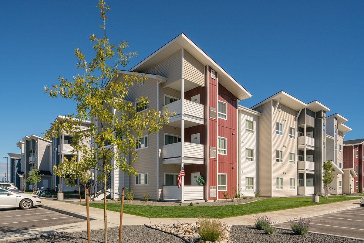 Modern apartment buildings with a mix of beige and red siding, featuring balconies. Green lawn and trees in the foreground, with a clear blue sky above. Parking spaces visible in front of the buildings.
