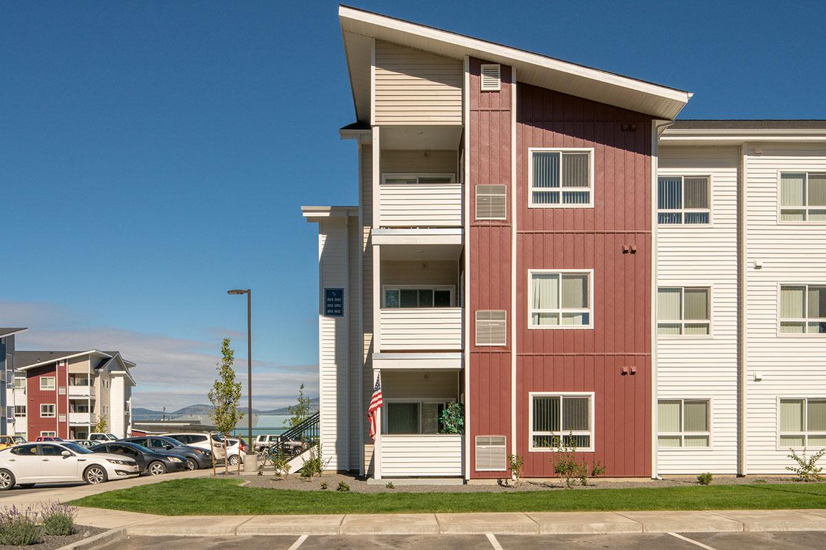 Three-story residential building with a combination of red and white siding, featuring multiple balconies and windows. An American flag is displayed on the ground level. The surrounding area includes parked cars and landscaped greenery under a clear blue sky.