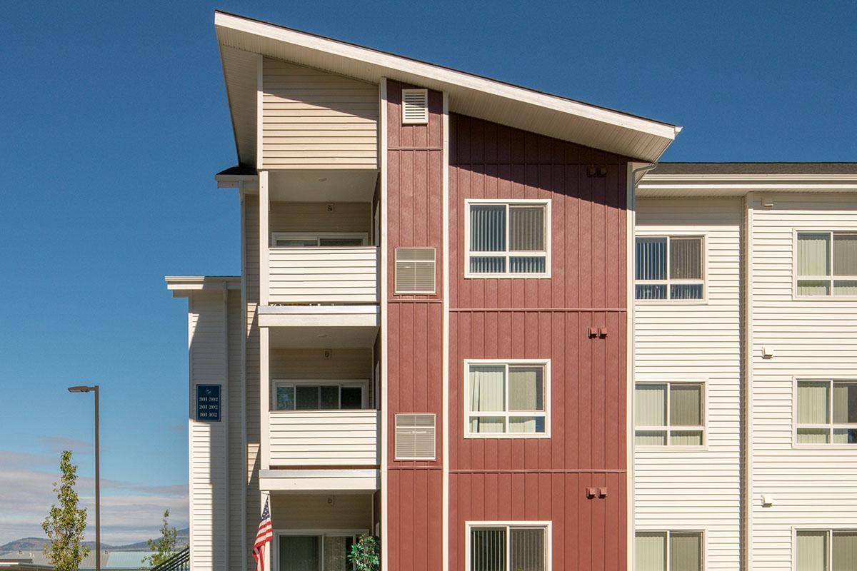 A modern three-story apartment building with a mix of red and white siding. The architecture features a sloped roof and balconies. An American flag is visible in front, along with trees and a clear blue sky in the background.