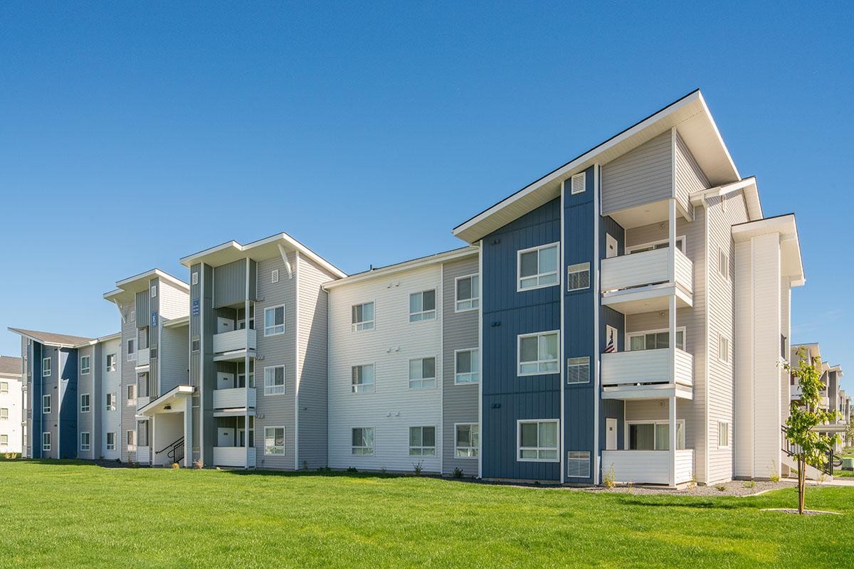 A modern apartment complex featuring two buildings with a mix of light gray and blue exteriors. The structure has three stories, balconies on some units, and is surrounded by green grass under a clear blue sky.