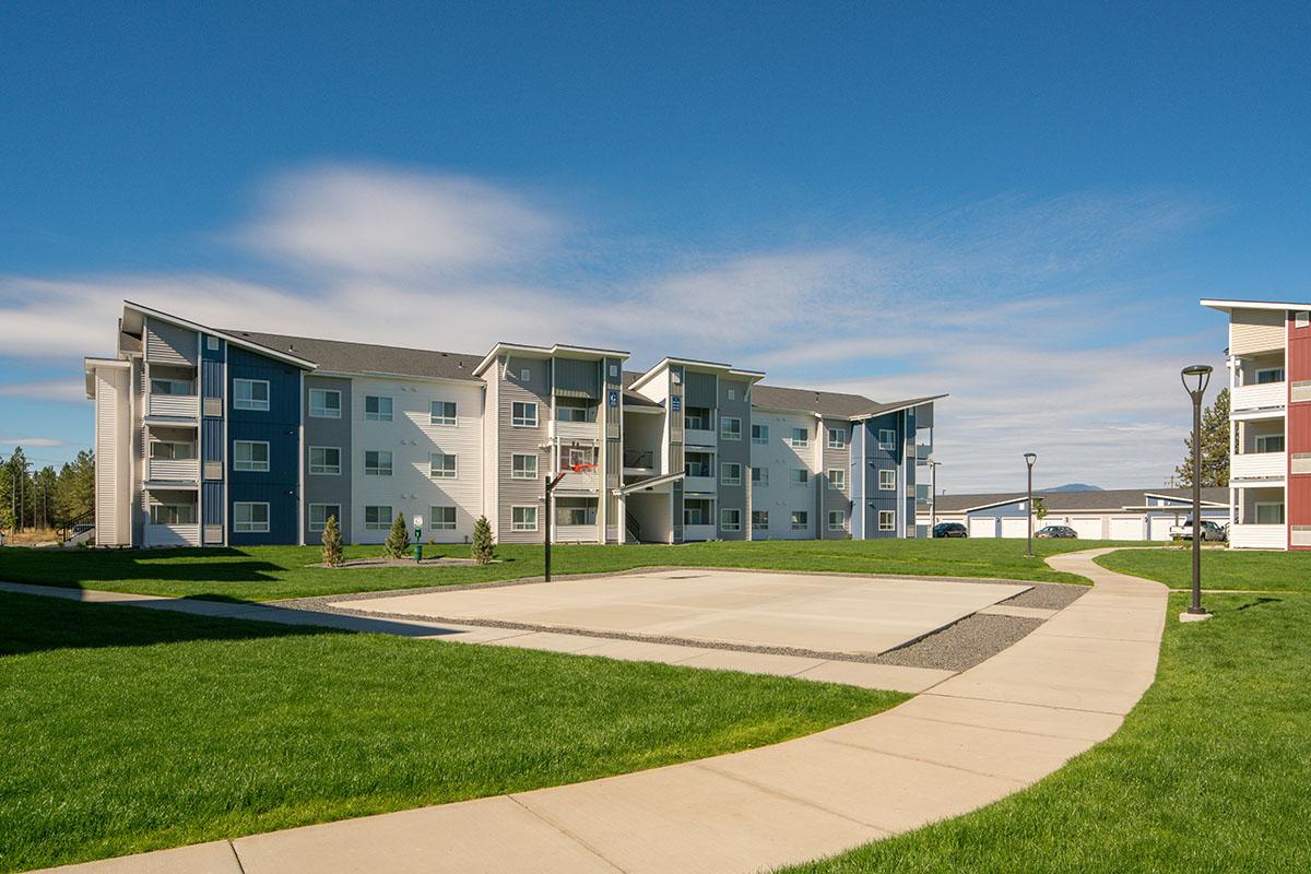 A modern apartment complex with a basketball court in the foreground, surrounded by green grass. The building features a mix of blue and white exterior colors. A clear blue sky serves as the backdrop, enhancing the bright and inviting atmosphere of the location.