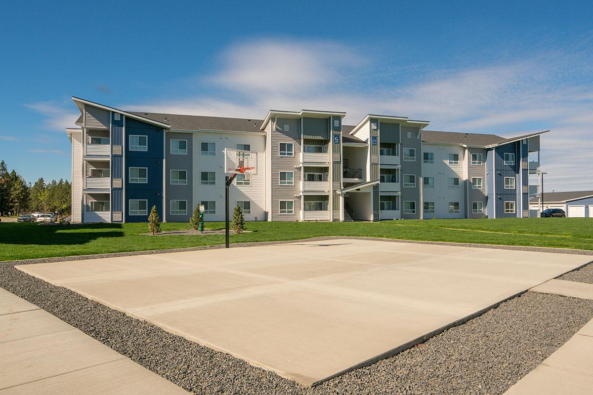A modern apartment building with multiple floors and balconies, set against a clear blue sky. In the foreground, there is a concrete basketball court with a hoop. The surrounding area features green grass and trees, creating a vibrant and inviting residential environment.