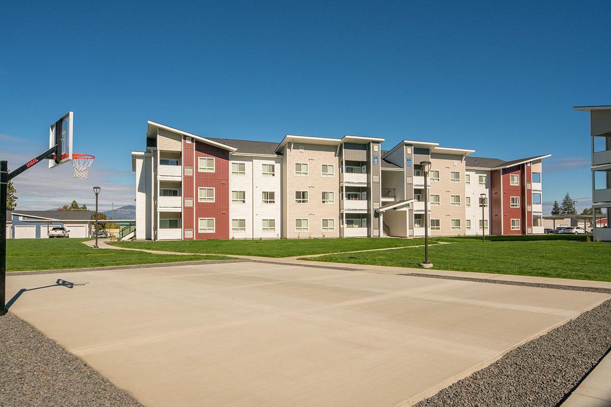 A modern apartment complex with three stories, featuring a mix of light and dark exterior colors. In front, a basketball hoop stands on a paved court, surrounded by well-maintained grass and pathways, under a clear blue sky.