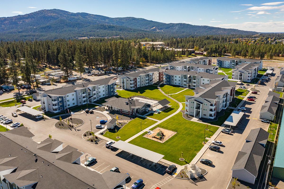 Aerial view of a residential complex featuring multiple buildings arranged around landscaped green spaces. The area includes parking, walking paths, and benches, with surrounding trees and mountains in the background under a clear blue sky.
