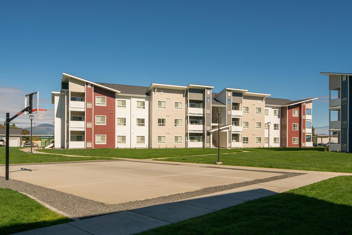 A modern apartment building with three stories, featuring a mix of beige and red exterior colors. In the foreground, there is a basketball court with a hoop and a concrete surface, surrounded by green grass and clear blue skies.