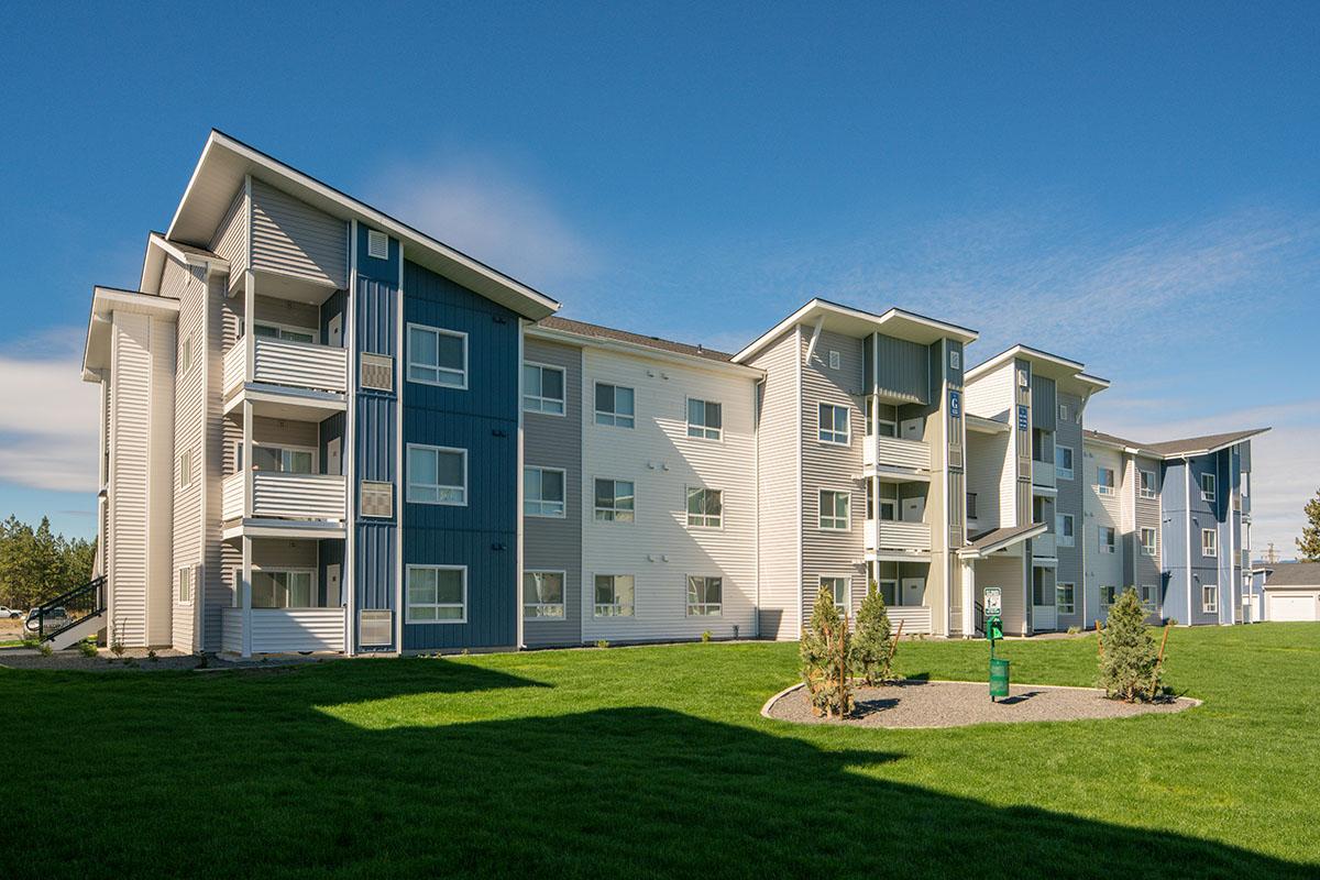 A modern three-story apartment building with a mix of blue and white siding. The building features multiple balconies and large windows, surrounded by green grass and a landscaped area. Clear blue sky above enhances the vibrant appearance of the property.