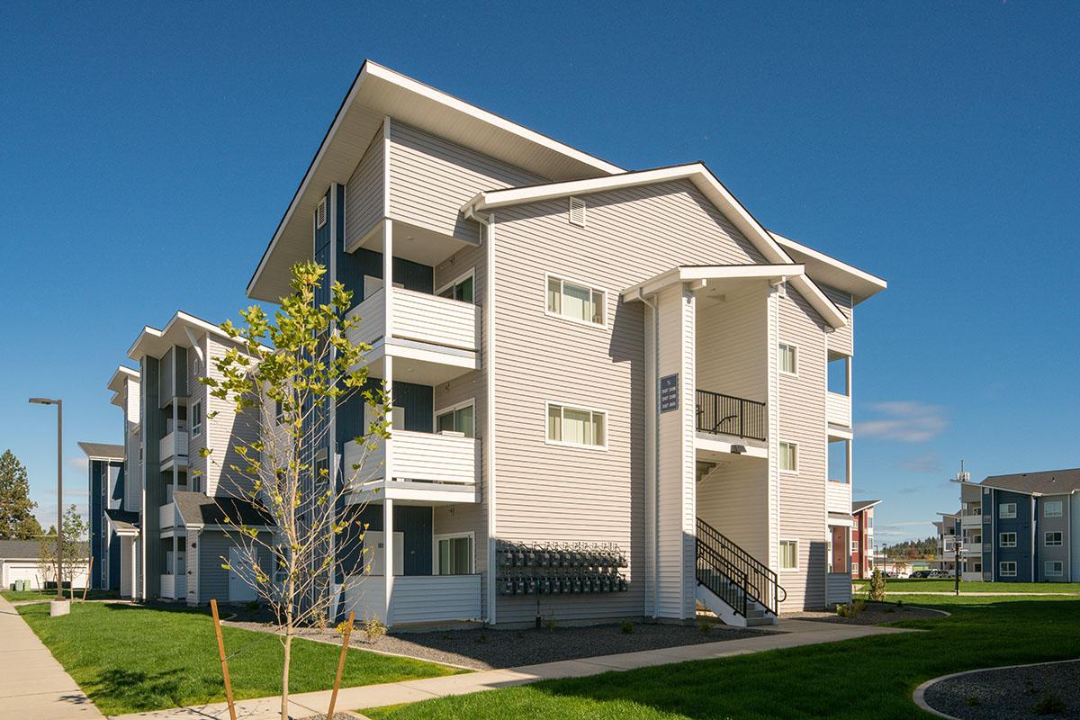 A modern multi-unit residential building with three levels, featuring a light-colored exterior and balconies. The foreground includes young trees and a pathway, while the background shows additional similar structures under a clear blue sky.