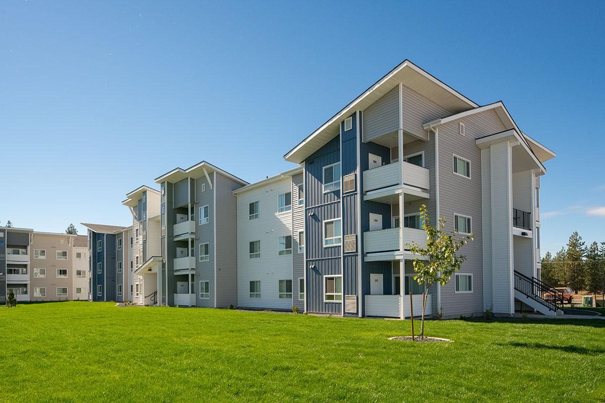 A modern apartment complex featuring several three-story residential buildings with balconies. The façade combines white and blue siding, and the surrounding area includes well-maintained green grass and small trees under a clear blue sky.