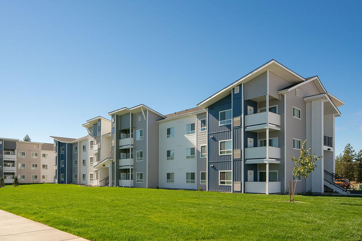 Modern multi-unit apartment building with a combination of white and blue siding. The structure features three stories, multiple balconies, and large windows. Surrounding the building is a well-maintained green lawn under a clear blue sky.
