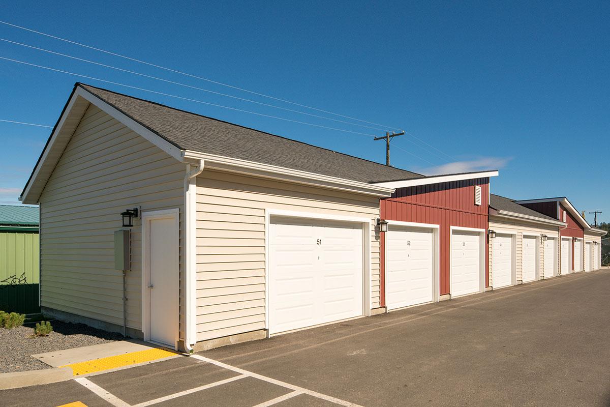 Row of storage units with beige and red exteriors, featuring white garage doors. The scene is set under a clear blue sky, with a paved driveway in front. Utility poles are visible in the background, and there is gravel landscaping alongside the units.