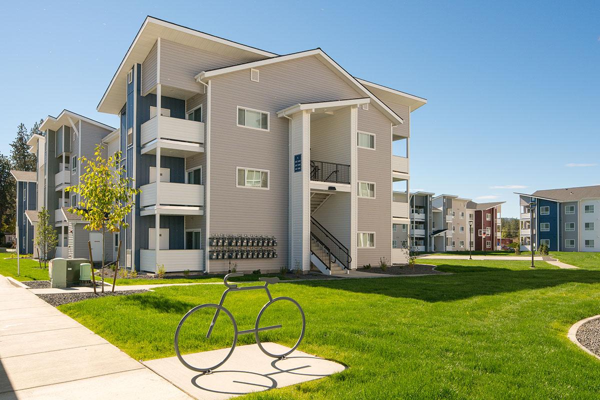 Modern apartment complex with light gray and blue buildings, featuring balconies and staircases. A metal bicycle sculpture sits on a grassy area, with walkways and landscaped grounds. Clear blue sky in the background.