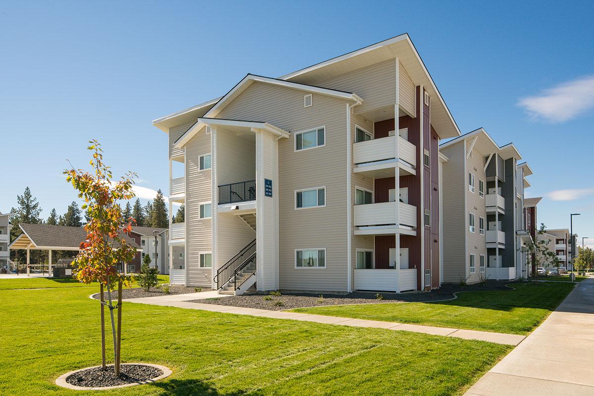 A modern three-story apartment building with a mix of beige and red siding, featuring multiple balconies and stairs. In the foreground, a small tree and well-maintained green lawn enhance the surroundings, with a sidewalk leading up to the building. Clear blue sky serves as the backdrop.