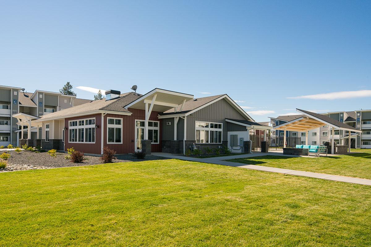 Modern residential building with a red and gray exterior, featuring large windows and a landscaped lawn. In the foreground, well-maintained grass and a paved pathway lead to the entrance. Sunlit sky overhead with a few clouds, and contemporary outdoor seating visible in the distance.