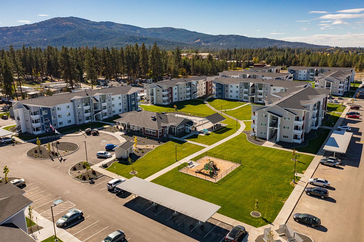 Aerial view of a residential complex featuring multiple buildings, green lawns, and parked cars. The area includes playground equipment and is surrounded by trees and mountains in the background, under a blue sky with scattered clouds.