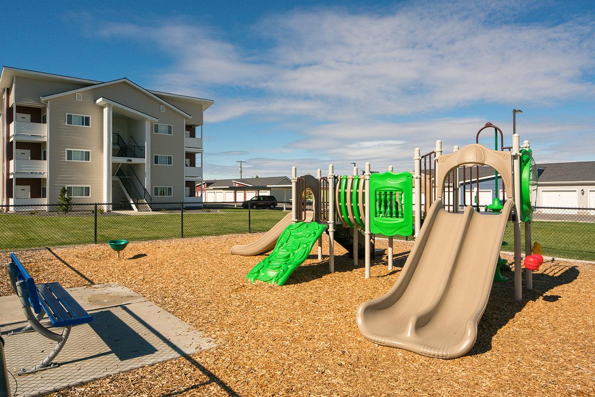 A colorful playground featuring green and tan slides, climbing structures, and safety features, situated on a bed of wood chips. Nearby is a blue bench and a grassy area with a fence. In the background, a multi-story apartment building and other structures can be seen under a clear blue sky.