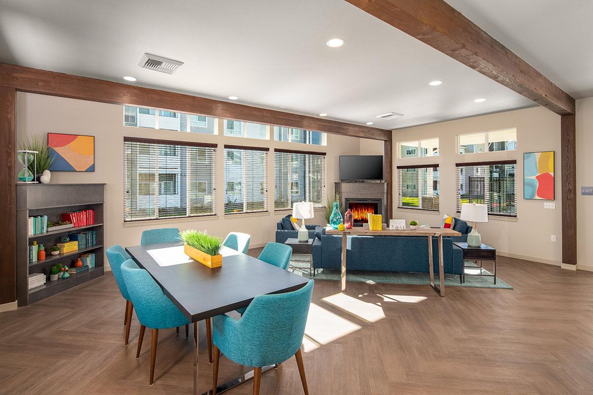 A well-lit modern lounge area featuring a dining table with turquoise chairs, a cozy seating arrangement with a fireplace, and large windows allowing natural light. Colorful abstract artwork adorns the walls, and plants add a touch of greenery to the space.
