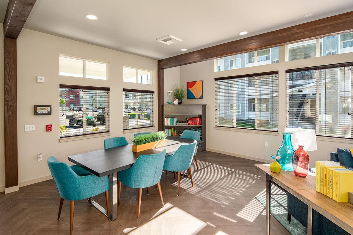 A modern common area featuring a dining table surrounded by teal chairs. There are large windows allowing natural light, plants on the table, colorful artwork on the walls, and a bookshelf filled with books. The decor includes a mix of wood and contemporary finishes, creating a welcoming atmosphere.
