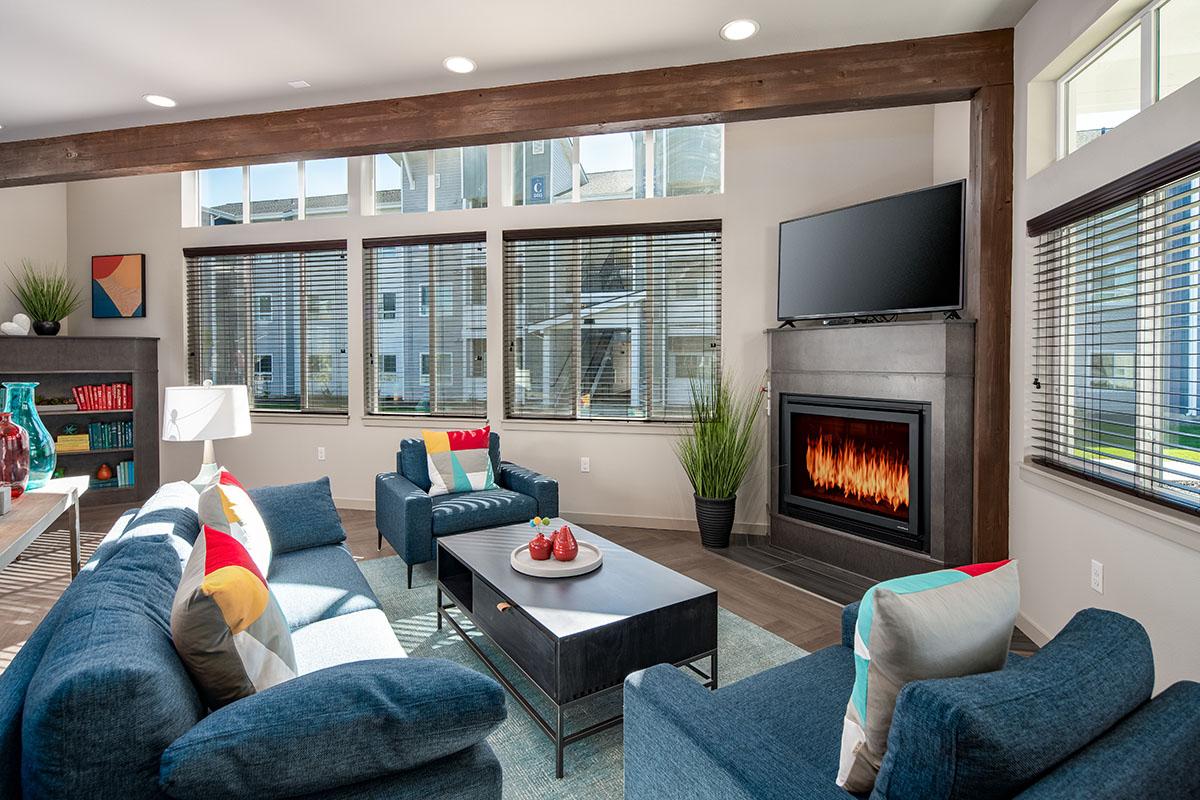 Cozy living room with modern furnishings, including a blue sofa set, a black coffee table, and a contemporary fireplace. Large windows let in natural light, framed by blinds. Decorative plants and colorful cushions add a vibrant touch to the inviting space.