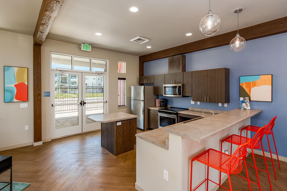 Modern kitchen with a central island, featuring sleek cabinetry and stainless steel appliances. Bright red bar stools line the counter. Large windows allow natural light to enter, and abstract paintings adorn the walls, creating a vibrant atmosphere. A door leads to an outdoor area.