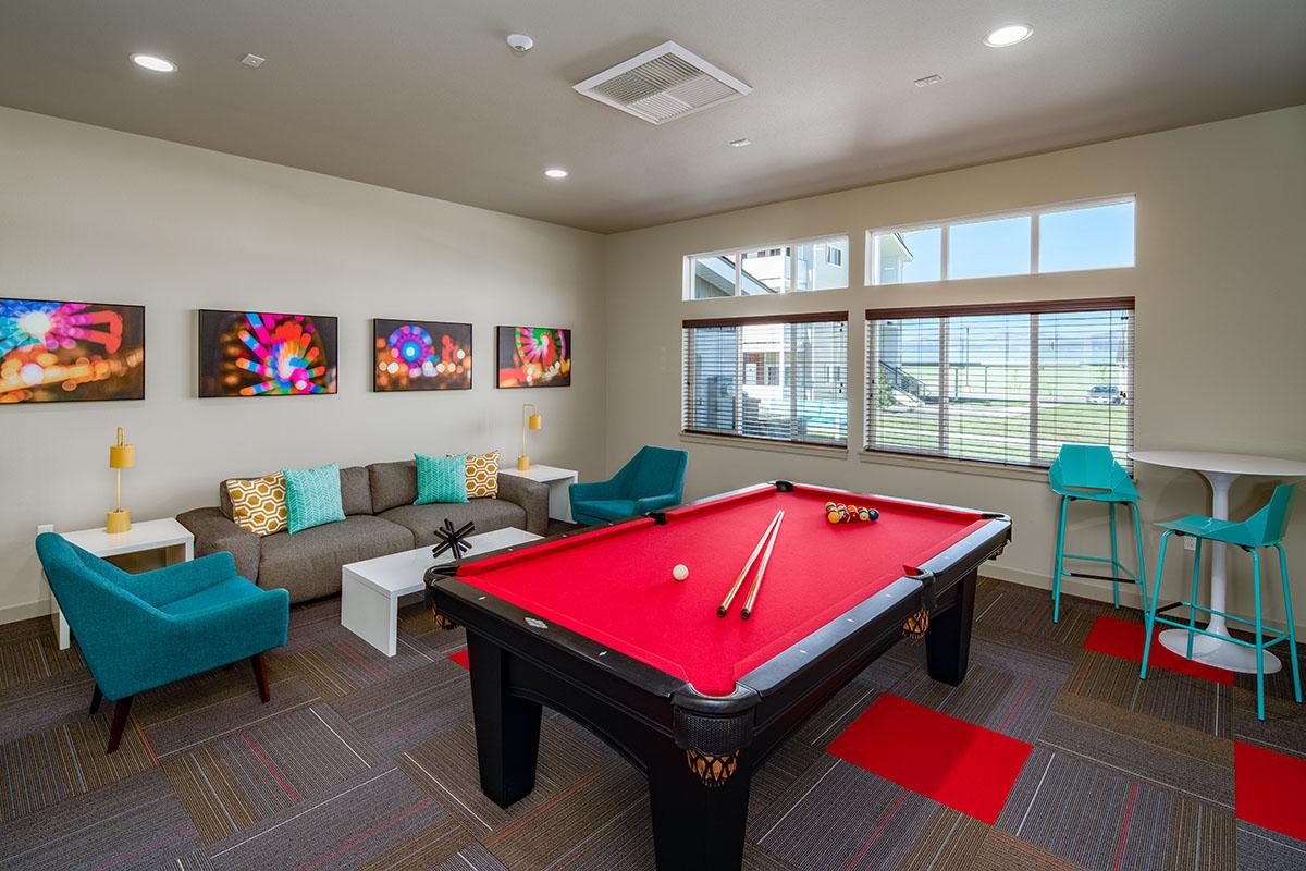 A bright and modern game room featuring a red pool table in the center, surrounded by comfortable seating. Stylish turquoise chairs and a cozy sofa with decorative pillows are positioned around, along with a small table. Large windows allow natural light to fill the space, and vibrant artwork adorns the walls.