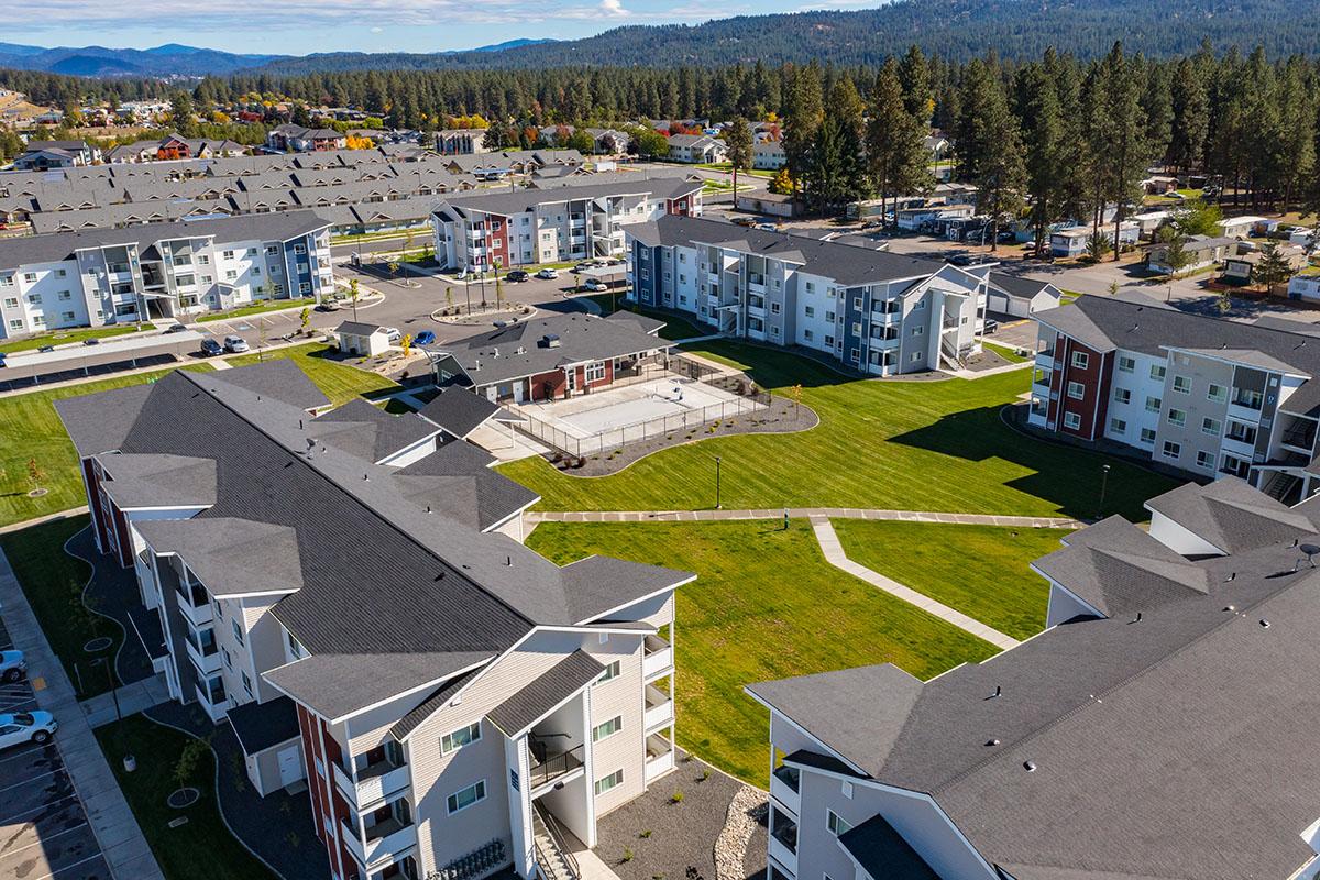 Aerial view of a modern residential complex featuring multiple apartment buildings with triangular roofs, surrounded by green lawns and landscaping. The area includes pathways and recreational spaces, with trees and a mountainous backdrop in the distance. The scene showcases a suburban environment.