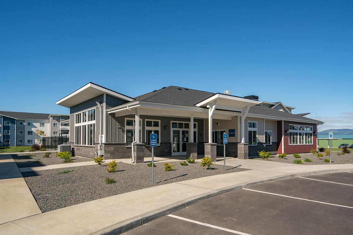 A modern single-story building with a sloped roof and large front windows, surrounded by a landscaped area. The facade features a mix of gray and red colors. A paved parking lot is visible in front, with designated accessible parking spaces. The sky is clear and blue in the background.