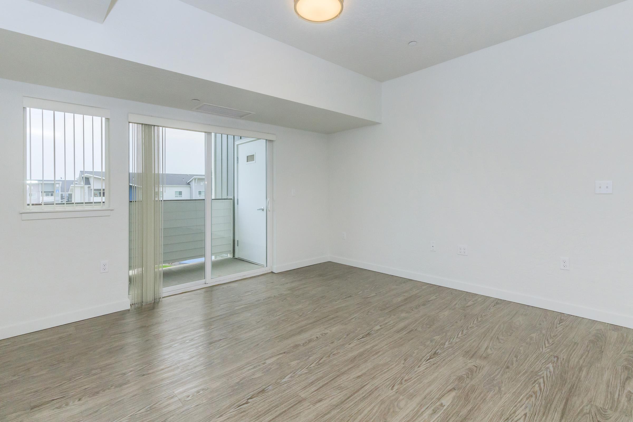 Empty room with light-colored walls and wooden flooring. There are large windows with vertical blinds, and a sliding glass door leading to a balcony. The ceiling has a round light fixture. The room appears bright and spacious, ideal for customization or furnishing.