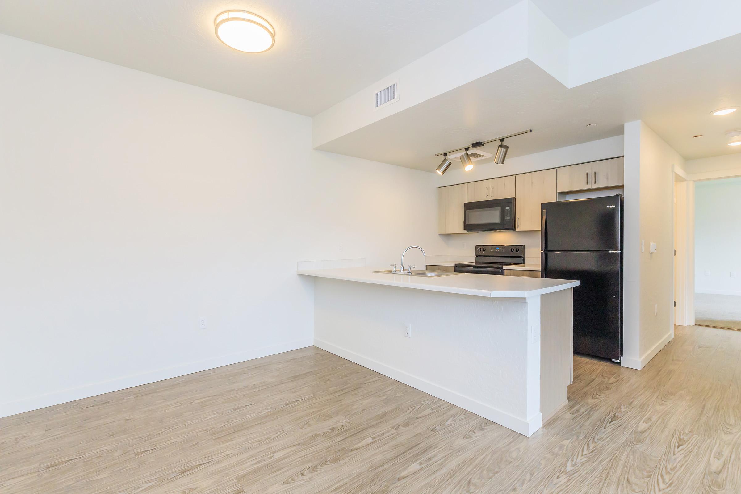A modern kitchen area featuring light-colored cabinetry, a stainless steel stove, microwave, and refrigerator. The kitchen island with a countertop separates it from the living space, showcasing a bright, open layout with light wood flooring and white walls.