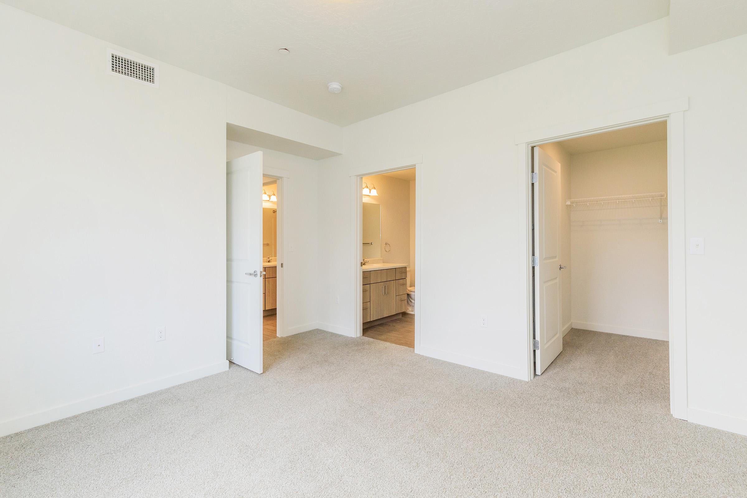 A bright, empty room with light-colored walls and carpet. Two open doorways lead to other areas, one potentially a bathroom and the other a closet. Natural light enters the space, emphasizing the clean and minimalist design.