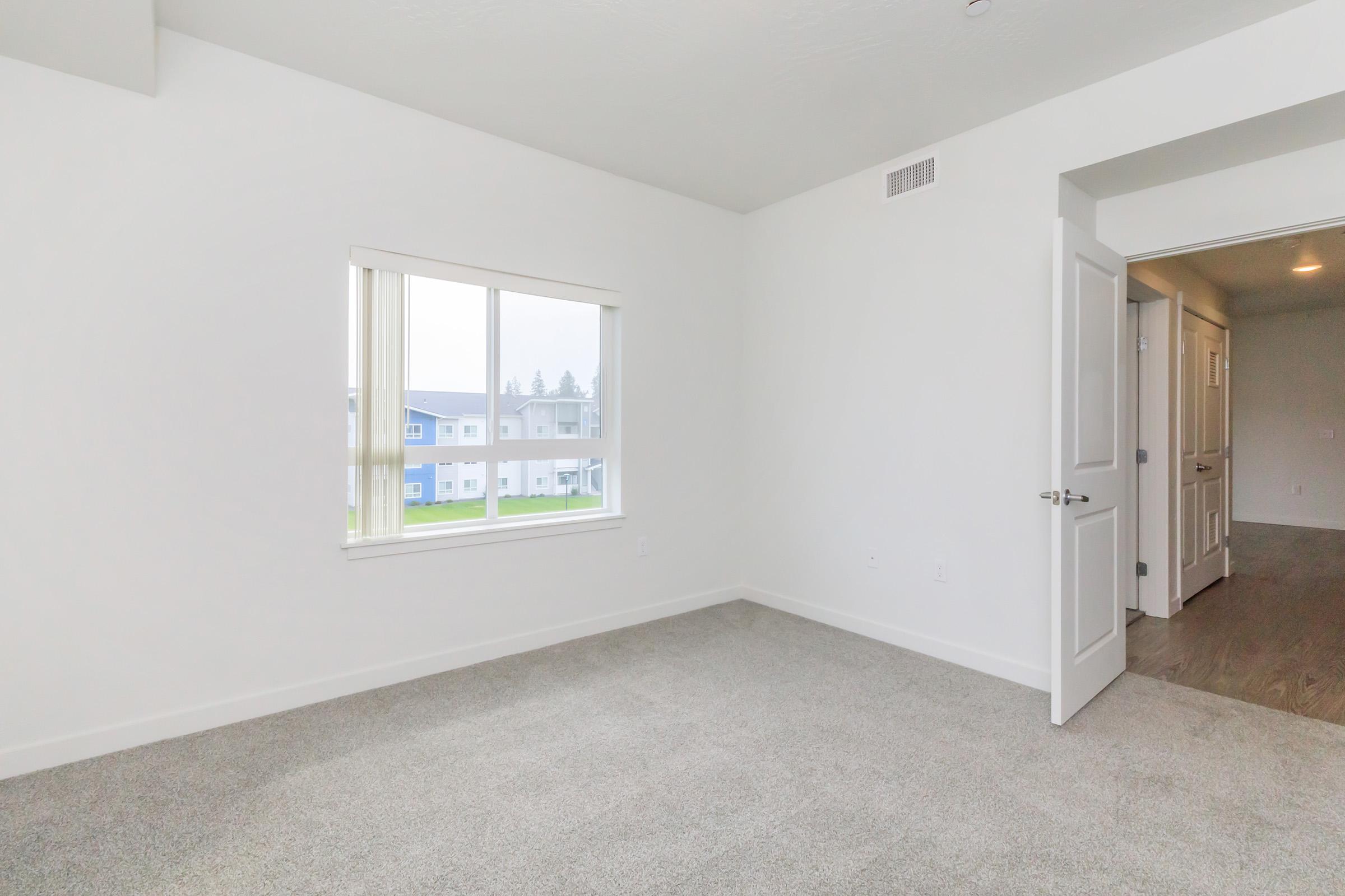 Empty room with light beige carpet and white walls. A large window lets in natural light, offering a view of a grassy area and other buildings outside. The room has a door leading to another space and is sparsely furnished, creating a spacious and minimalistic atmosphere.