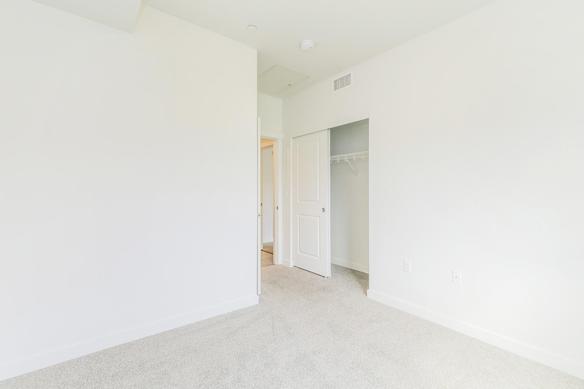 A bright, empty room with white walls and carpeted floor. There is a doorway leading to another room on the left and a closet with sliding doors on the right. Natural light fills the space, creating an airy atmosphere, ideal for interior design or decorating ideas.